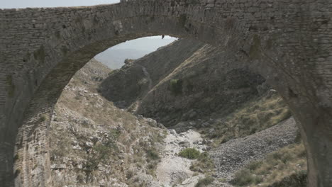 Drone-shot-of-the-Ali-Pasha-Bridge-Outside-of-Gjirokaster,-Albania-on-a-sunny-day-sitting-in-between-the-rocky-mountains-with-a-dry-river-flying-underneath-to-reveal-the-city-and-landscape-LOG