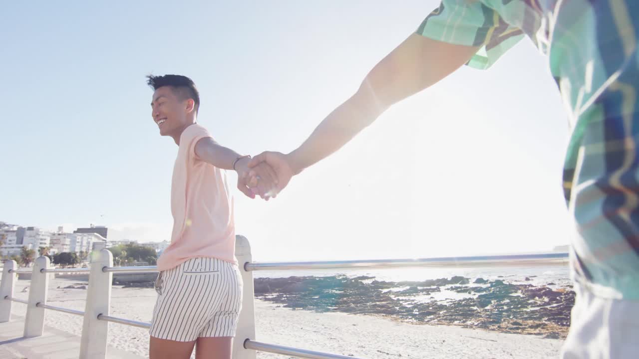 해변의 산책로에서 손을 잡고  ⁇ 고 있는 행복한 다양한 동성애 남성 커플, 느린 동작