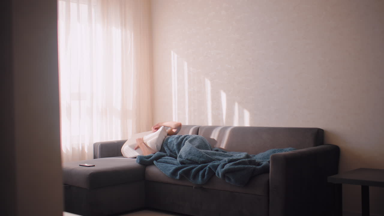 Young woman wrapped in fluffy blue blanket lying on couch, hiding face with pillow to block out bright morning sunlight, peaceful shadows cast through sheer curtain onto wall