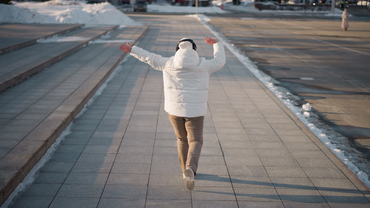 Back view of youth grooving down tiled walkway beside steps on busy winter street lined with snow piles, wearing puffy white jacket and beanie, surrounded by billboards, and city buildings