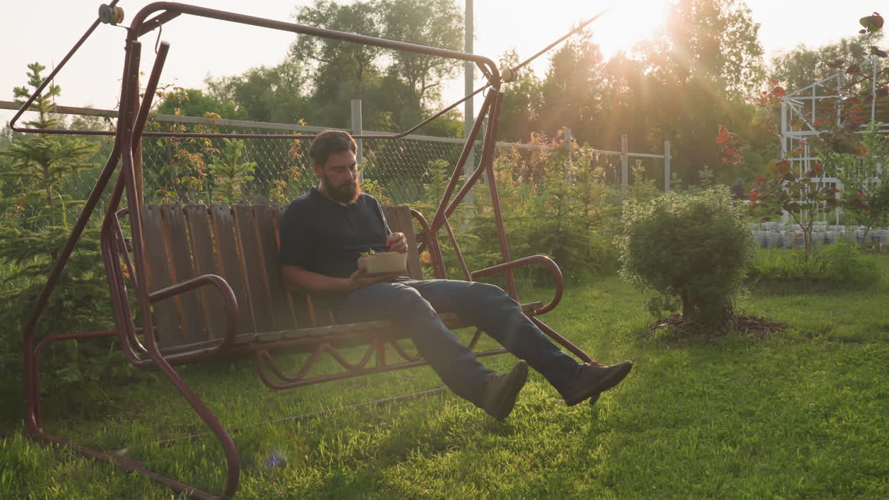 man seated on wooden swing bench in home garden gently swinging while eating ripe freshly harvested strawberry from biodegradable carton under warm golden sunset glow with lush foliage
