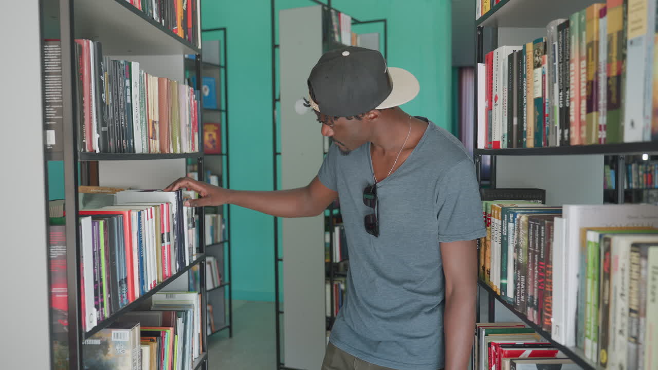 Hombre negro con ropa informal y gafas de sol colgando de la camisa caminando entre las estanterías de una biblioteca, hojeando títulos en un entorno académico tranquilo con una pared color verde azulado, centrado en explorar la literatura.