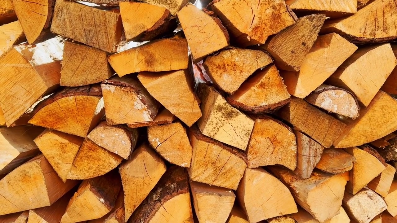 Neatly stacked split firewood in bright sunlight, showing golden log faces, bark edges, varied shapes, and dry household fuel ready for heating.