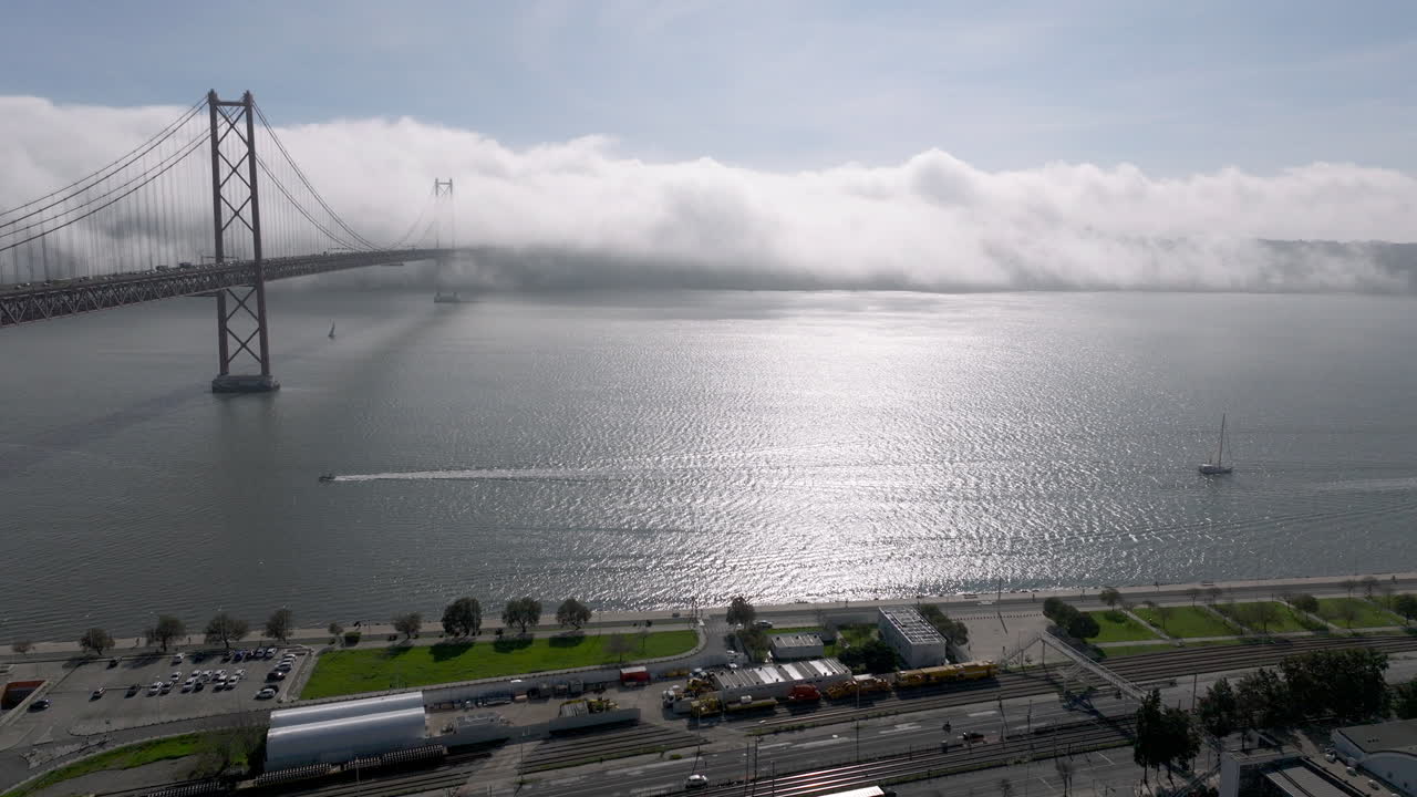 Aerial drone shot towards the 25th April Bridge and waterfront in Lisbon, Portugal, Europe. Sunny and bright with clouds, fog, mist covering the bridge. Shot in ProRes 422 HQ
