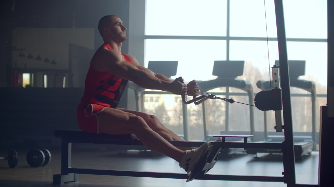 A man pulls weight to his stomach sitting in the simulator performing an exercise for the muscles of the back against the background of large Windows of the gym. Powerful male athlete doing rowing exercises