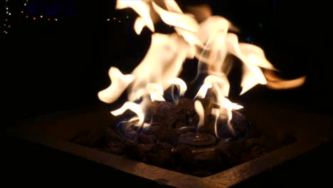 Full-frame-shot-of-outdoor-fireplace-burning-with-people-walking-by-in-the-shadows-in-front-of-twinkling-Christmas-lights