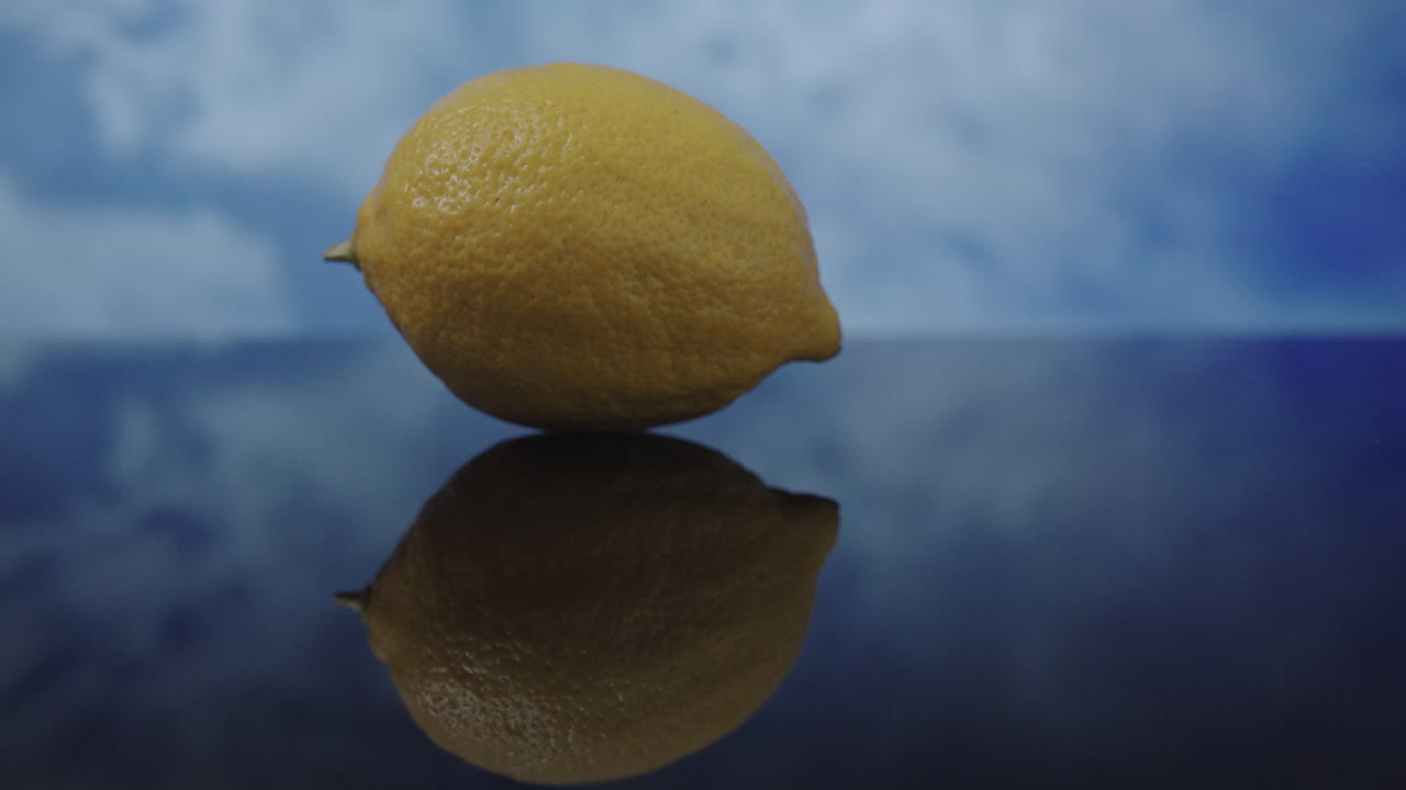 Un limone giallo appoggiato su una superficie asciutta e riflettente, mentre le nuvole si muovono velocemente sullo sfondo durante un time-lapse in una giornata nuvolosa