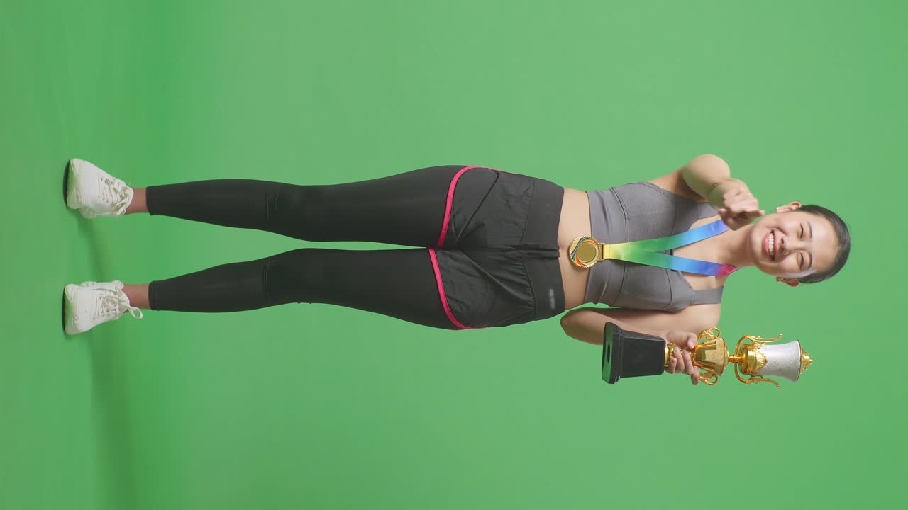 Full Body Of Asian Woman With A Gold Medal And Trophy Touching Her Chest Then Smiling And Pointing To Camera Being Proud Of Herself On Green Screen Background In The Studio