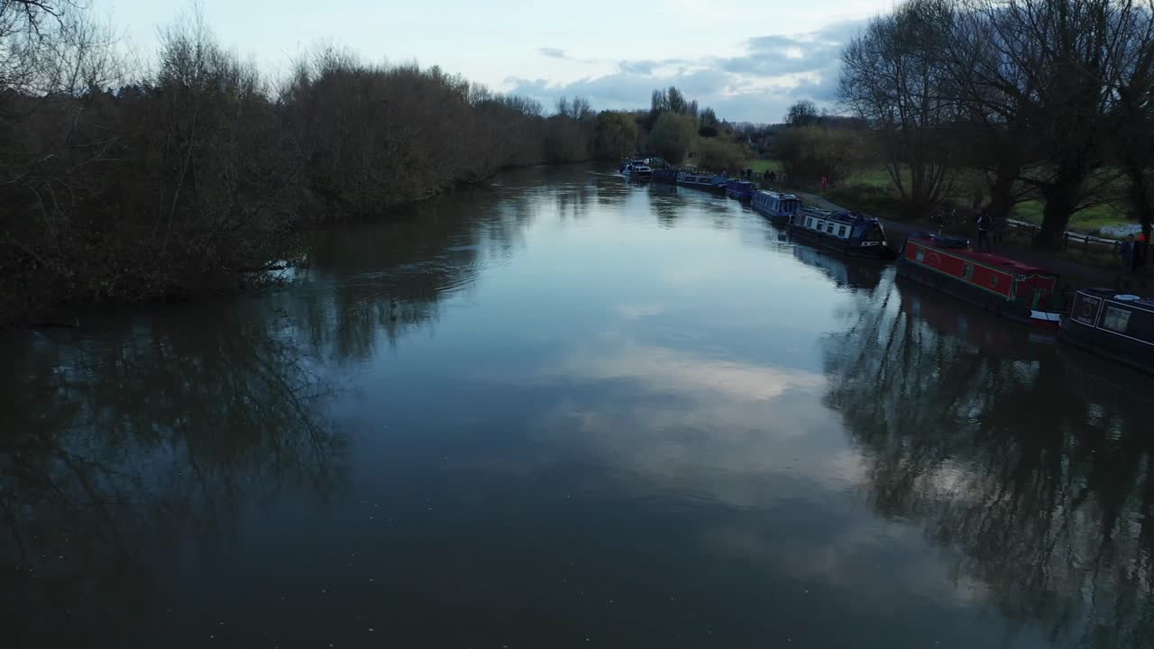 People walk along the shores of The Isis river (Thames) in Oxford. Boats docked on shore. Dolly in aerial flight over the river, with trees reflected in the water