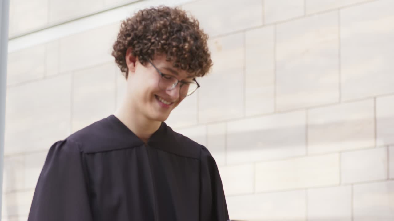 Formado do sexo masculino vestindo beca de formatura e lendo mensagem comemorativa enviada no smartphone do lado de fora do campus, sorrindo