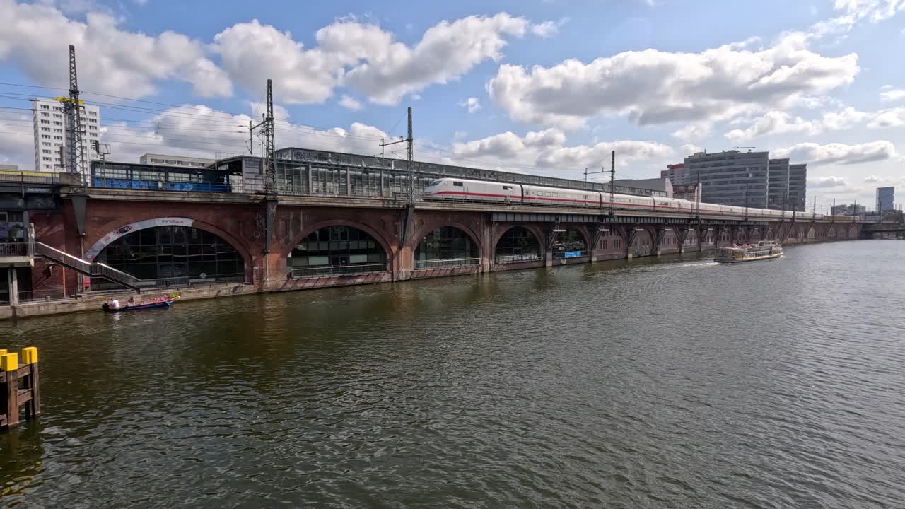 A passenger train travels across a historic brick railway bridge spanning a river in central Berlin, with a boat cruising below on a sunny day. Wide, steady shot with natural daylight and urban waterfront backdrop