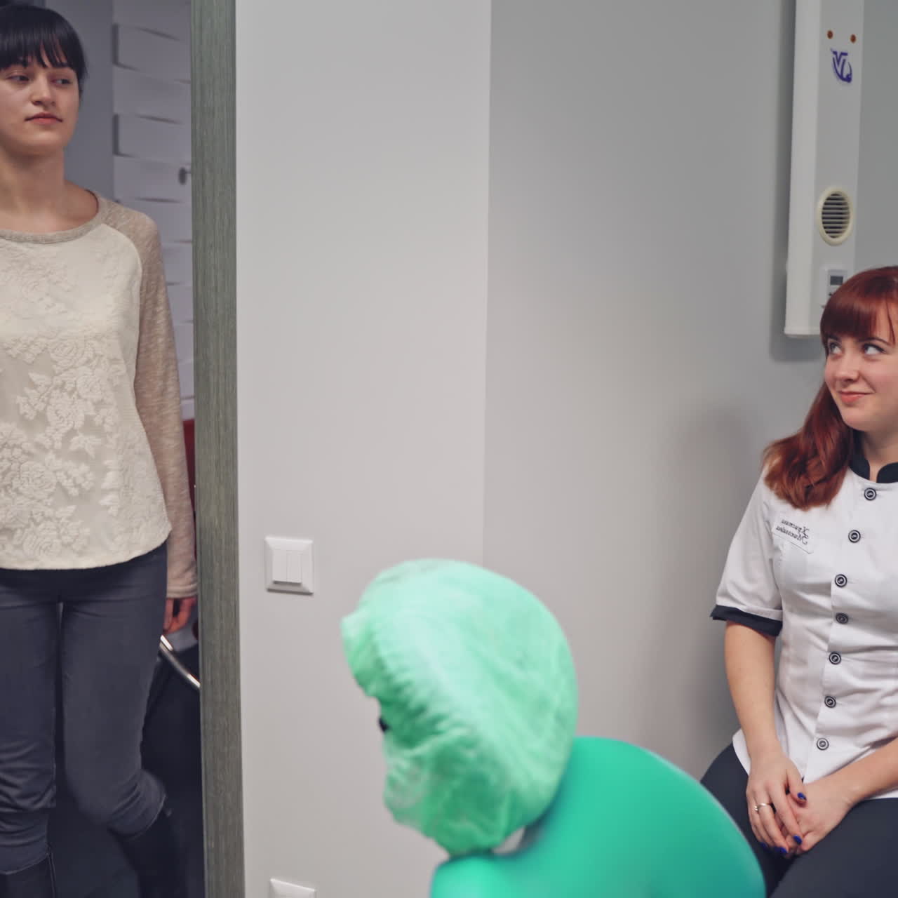 Young woman opens the door and comes to a dental office for check up her teeth. Female dentist is waiting for patient and showing to sit to the dentist chair
