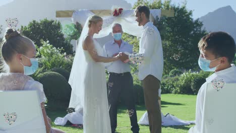 caucasian boy and girl in face masks watching happy couple getting married outdoors