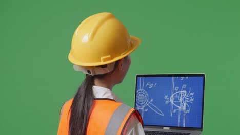 vista posterior cercana de una ingeniera asiática con casco de seguridad mirando el plano de la turbina eólica en una computadora portátil mientras está de pie en el estudio de fondo de la pantalla verde
