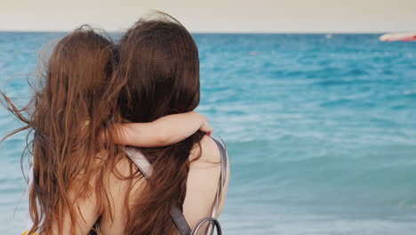 una joven madre con el pelo castaño abraza a su pequeña hija con el pelo largo y ondulado contra el mar azul