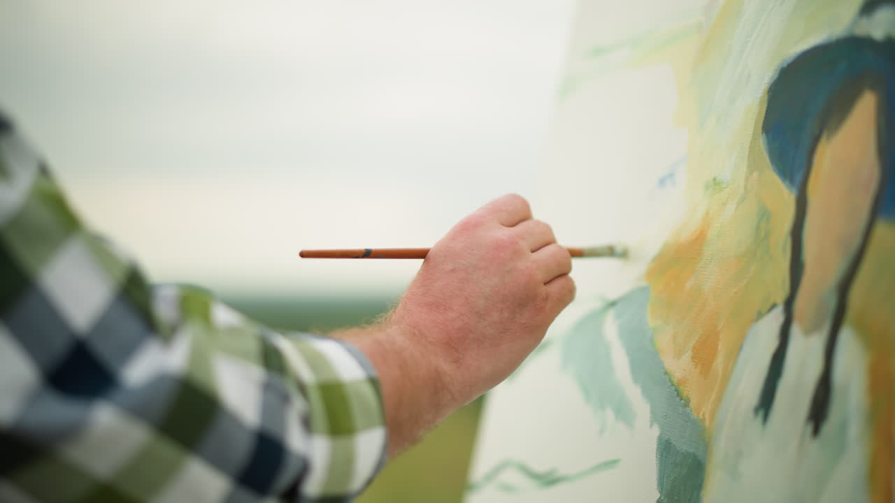 A close-up shot of an artist s hand in a checkered shirt holding a brush as he skillfully and gently paints a vibrant portrait on a canvas
