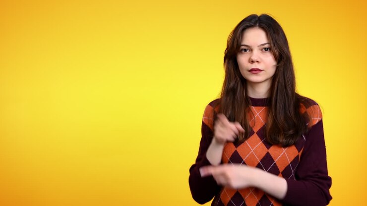 Giovane donna seria che indica qualcosa accanto a sé e mostra un gesto di rifiuto su sfondo giallo