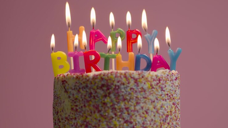 Lit Candles Spelling Happy Birthday On Cake Covered With Decorations Against Pink Studio Background