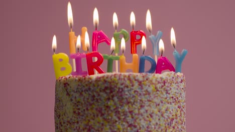 lit candles spelling happy birthday on cake covered with decorations against pink studio background