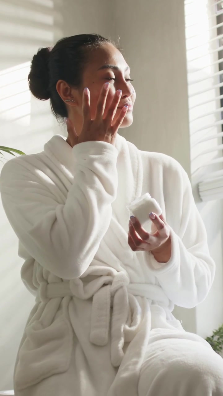 Vertical video: Holding jar, Asian woman scooping cream, rubbing eye for skincare at vanity in robe