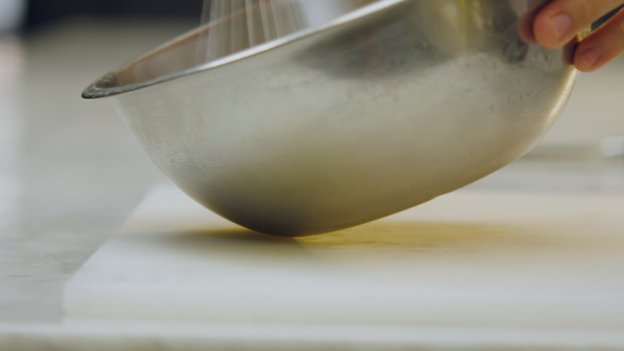 Close-up of a chef's hands beating a mixture with a hand whisk in an aluminum mold, placed on a chopping board on a kitchen counter