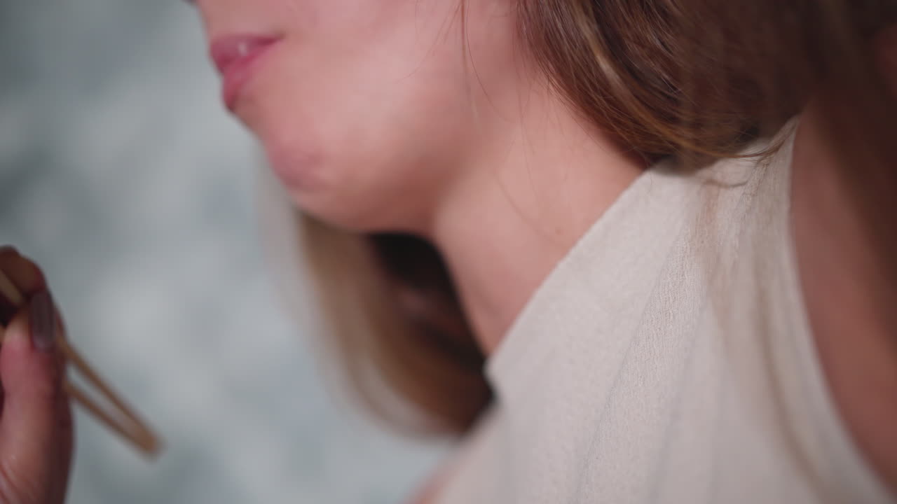 Close up of young woman eating sushi with chopstick as piece of sushi drops while chewing in profile view showing lower face neck and soft focus background
