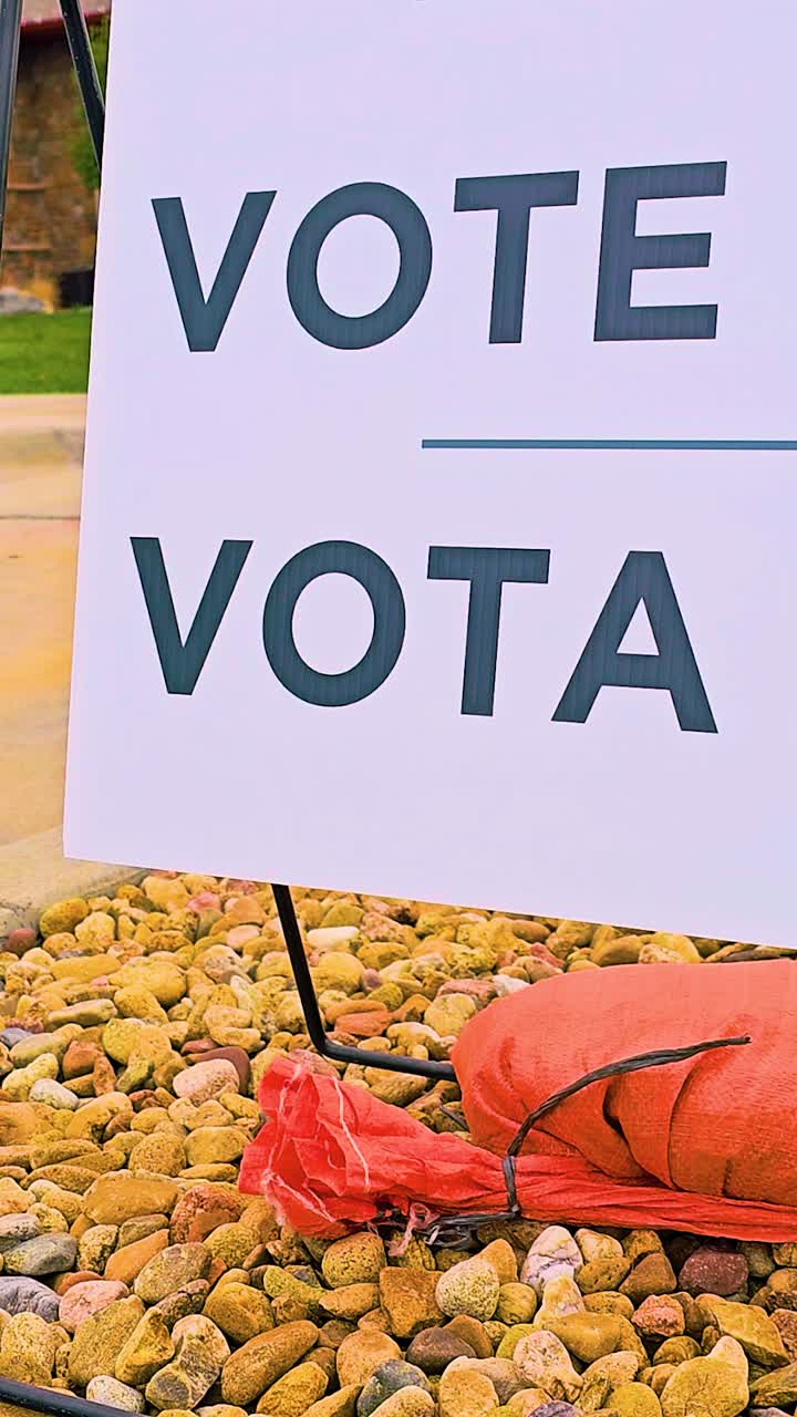 Close-up of a bilingual yard sign reading VOTE and VOTA on gravel with a sandbag, shot outdoors in daylight. Perfect for election, voter awareness, and civic engagement themes. Vertical Video