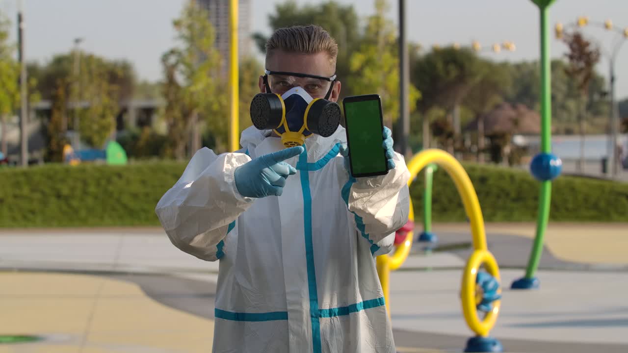 retrato hombre joven en un traje de protección, gafas, un respirador y guantes sostiene un teléfono inteligente con pantalla verde y tecla de croma con posición vertical de la pantalla y señala con un dedo.