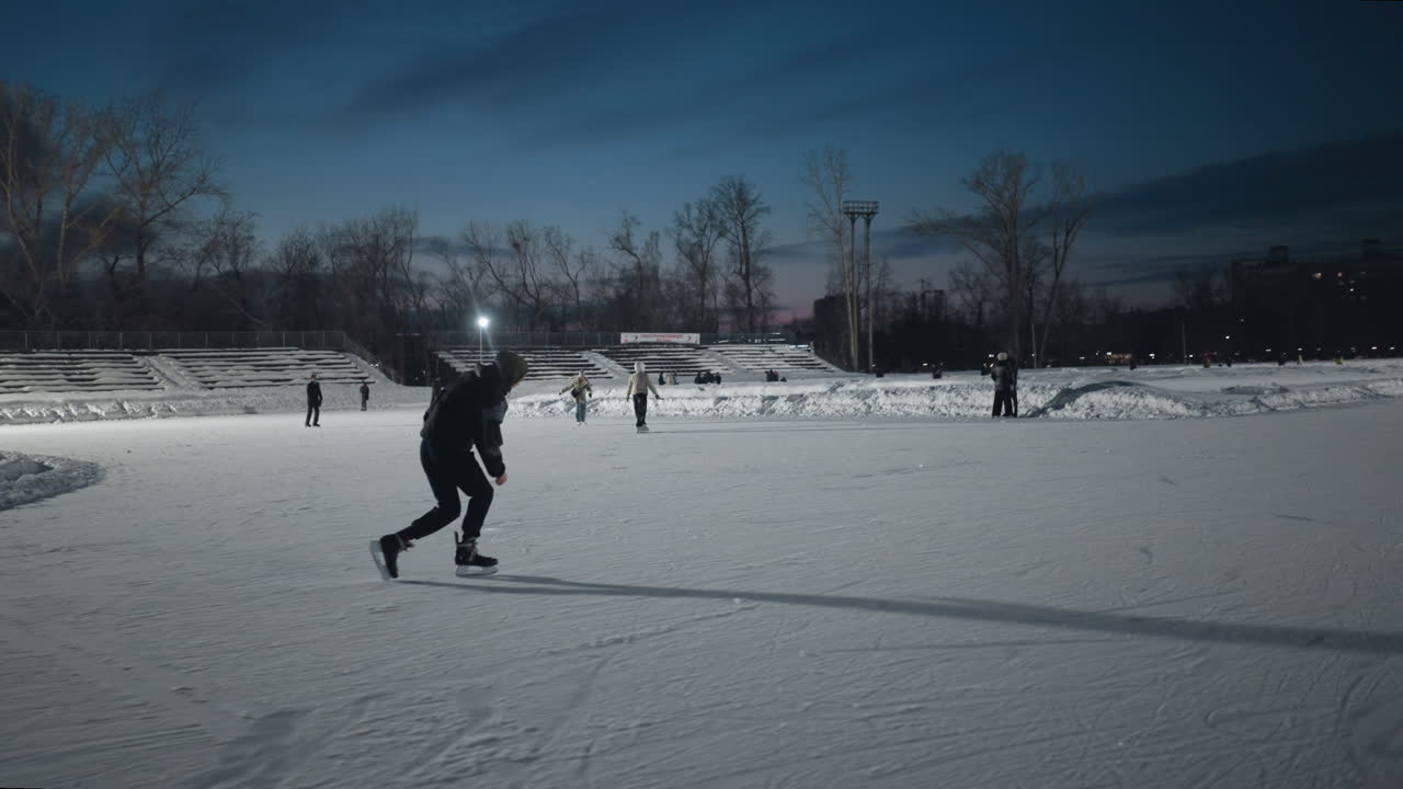Skaters gliding on smooth outdoor ice rink under evening sky with distant urban background and bright stadium lights illuminating rink while silhouettes of others skate peacefully in distance