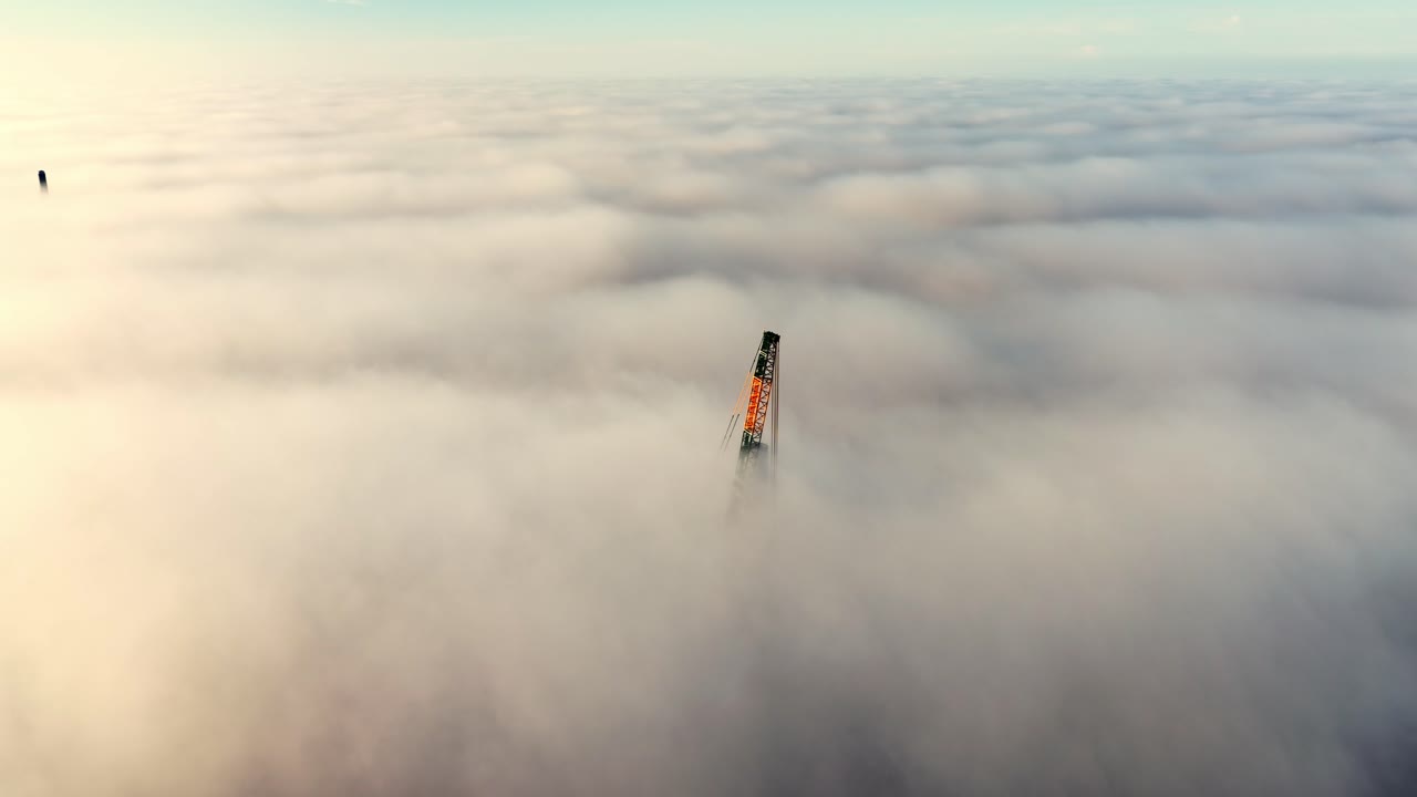 Construction crane arm emerging through dense morning fog during sunrise, aerial overview