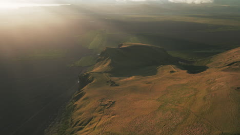 retirada aérea lenta sobre el acantilado dorado cerca de la playa de arena negra al atardecer, islandia