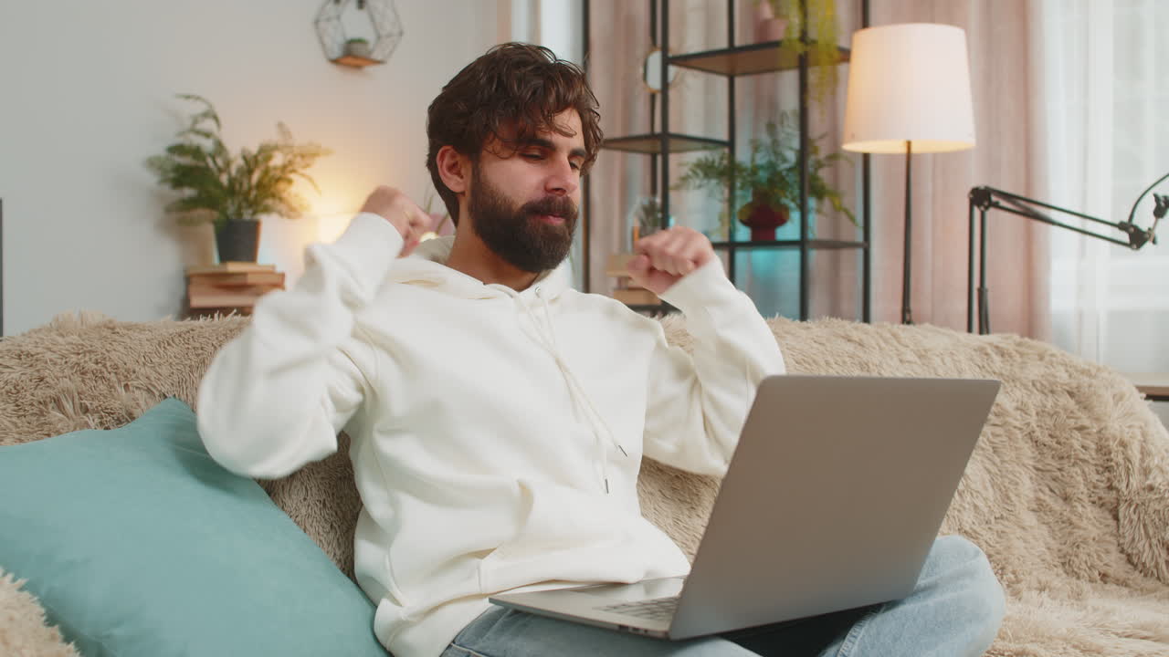 Excited young indian man freelancer using laptop computer sitting on sofa couch at home apartment