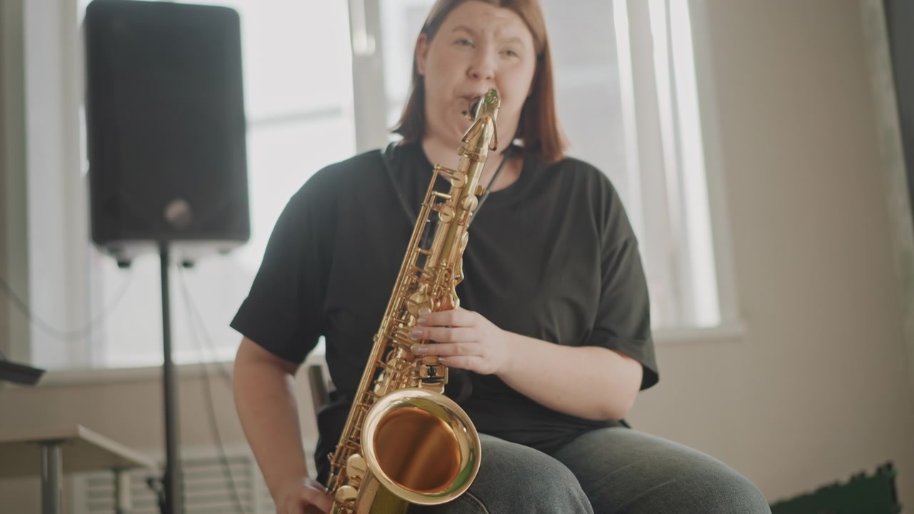 Gamba di musicista e primo piano di sassofono durante una dimostrazione di tecnica, l'insegnante si concentra sulla diteggiatura e sulla postura su uno sgabello di legno, tappeto testurizzato e amplificatore visibili, atmosfera didattica calma