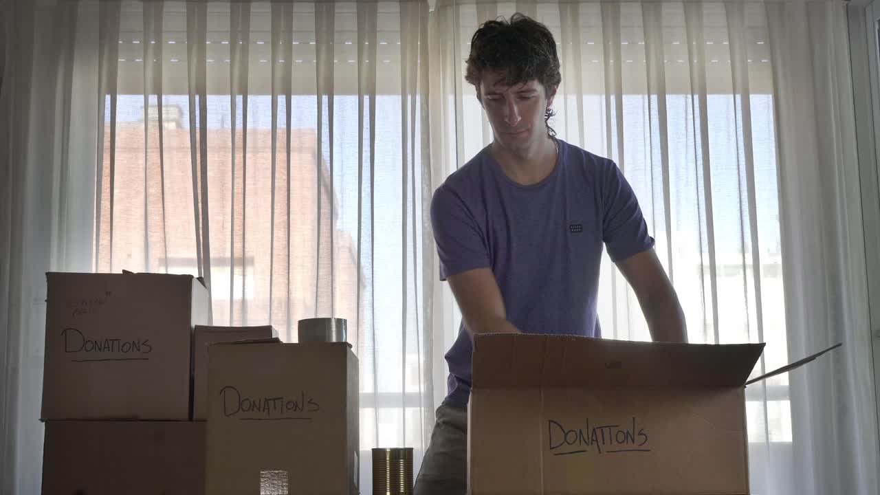 Young Man Packing Donation Box with Canned Goods and Giving Thumbs Up