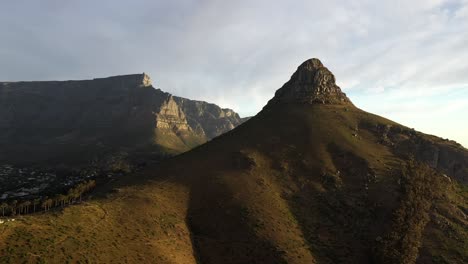 Toma-Giratoria-Aérea-Cinematográfica-Del-Pico-De-La-Cabeza-De-León-De-Ciudad-Del-Cabo-Con-La-Montaña-De-La-Mesa-Y-La-Colina-De-La-Señal-Durante-La-Puesta-De-Sol-De-La-Hora-Dorada