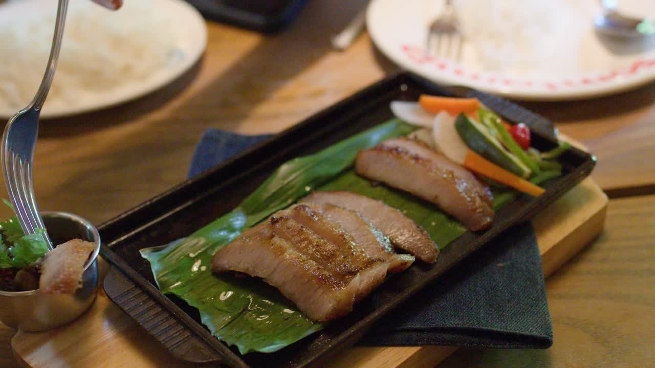 Hand dips grilled pork slice in spicy sauce with fork on wooden table, natural lighting