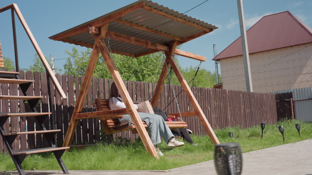 Two people enjoy sunny weather in backyard with wooden swing set, one sits reading on bench swing, other plays on round swing, cozy garden scene with wooden fence, grass, outdoor stairs