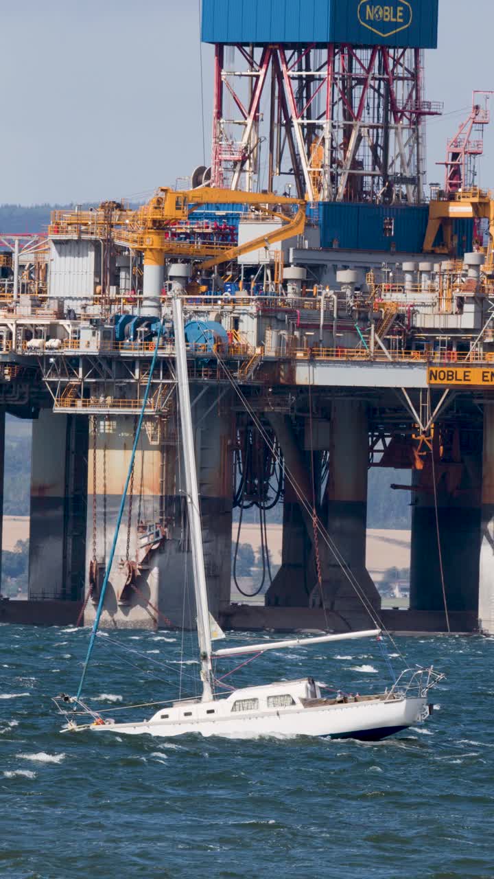 En hvid sejlbåd sejler forbi en stor offshore olieplatform i oprørt vand i dagslys, set med teleperspektiv.