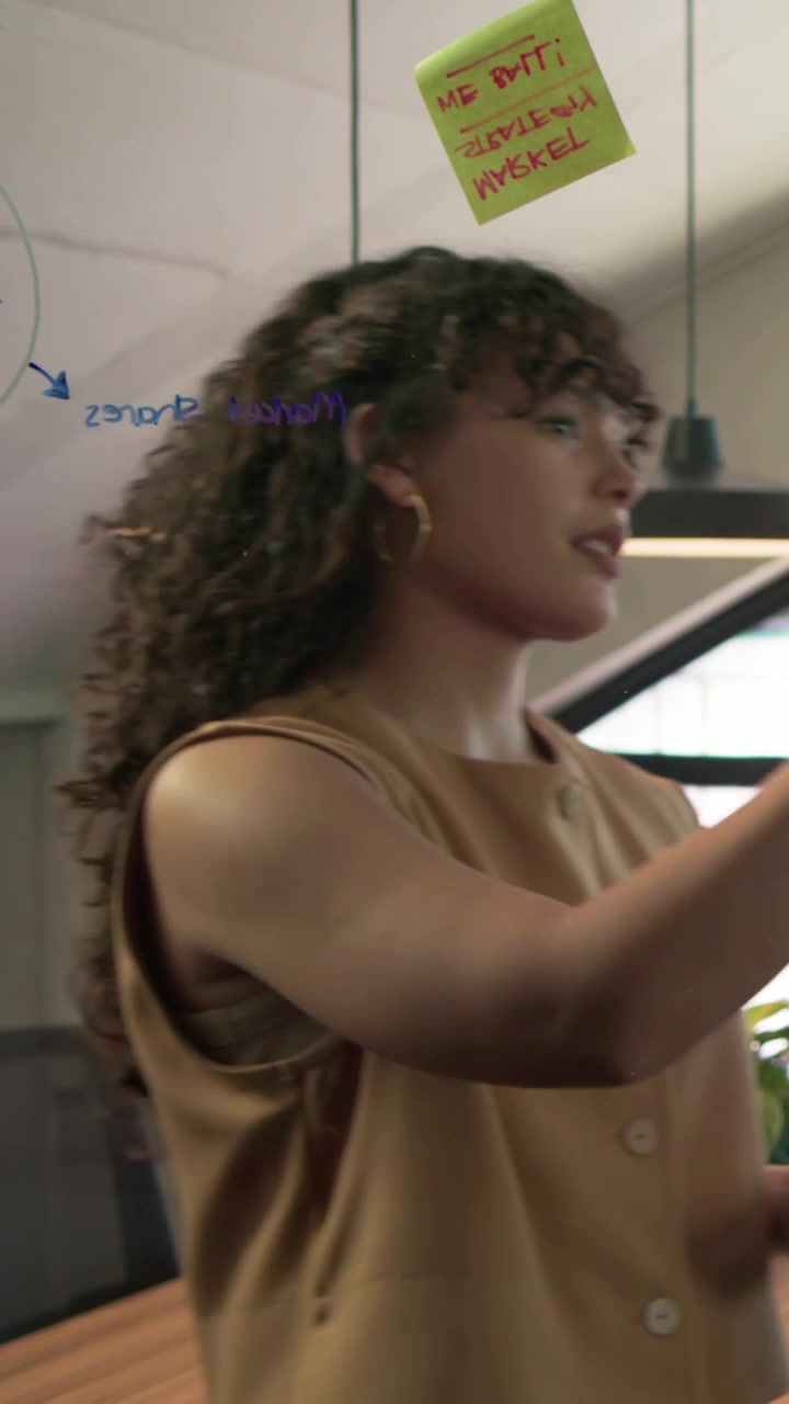Vertical video: Meeting starting, woman in tan top placing notes on glass board with marker to plan