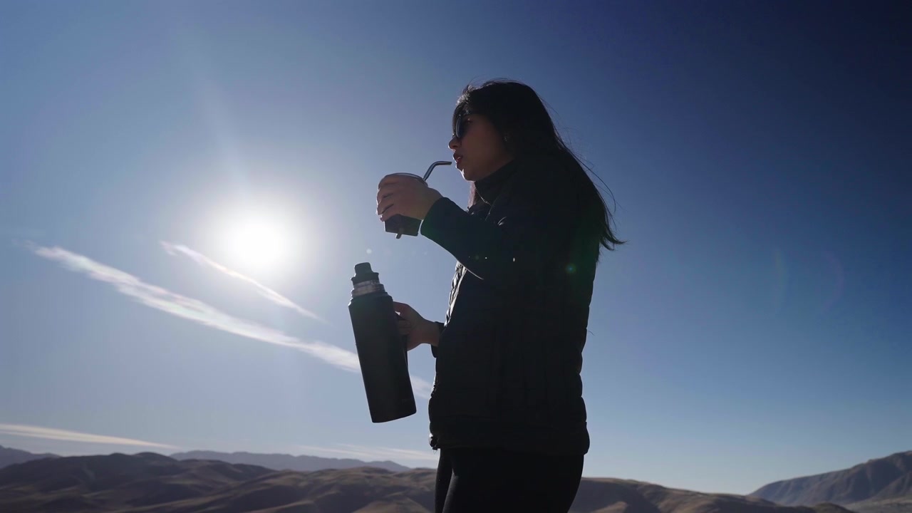 Donna che beve yerba mate a Tafí del Valle, Tucumán, Argentina, con la silhouette stagliata contro l'alba in montagna, con un tradizionale thermos in acciaio inossidabile e uno sfondo di paesaggio andino, ripresa al rallentatore