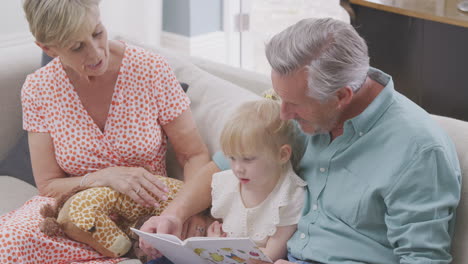 Abuelos-Sentados-En-El-Sofá-Con-Su-Nieta-En-Casa-Leyendo-Un-Libro-Juntos