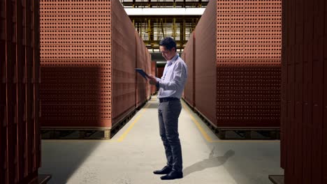full body side view of an asian male professional worker standing with his tablet with red brick packed in stacks are stored, working continuously with his tablet