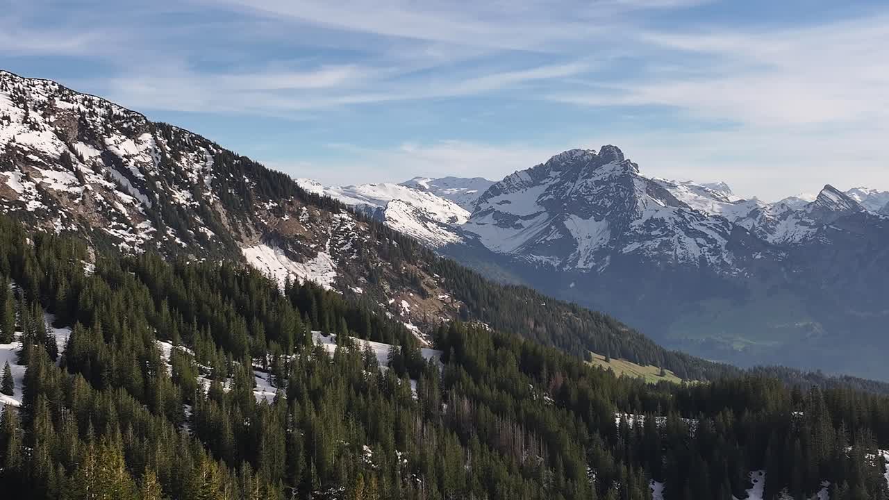 Stunning drone shot of Amden Arvenbuel, Switzerland, showing majestic snow-capped mountain ranges and picturesque canopies of alpine trees with blue skies and white clouds in a winter day