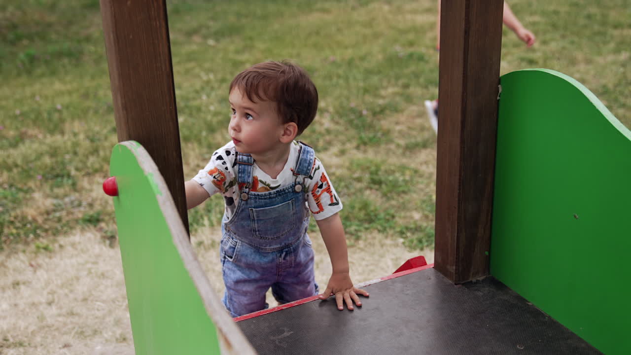 Charming baby boy climbing by the wooden steps to the slide. Toddler playing on the playground in summer. View from top.