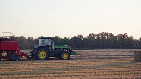 agricultural machine pressing hay on the golden field. harvesting dry grass for livestock. green and red tractor during seasonal works in summer.