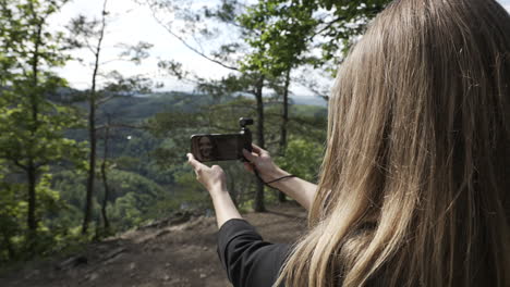 parte posterior de una mujer joven grabando vídeo selfie con una pequeña cámara cardán y un teléfono inteligente en el bosque en un día soleado