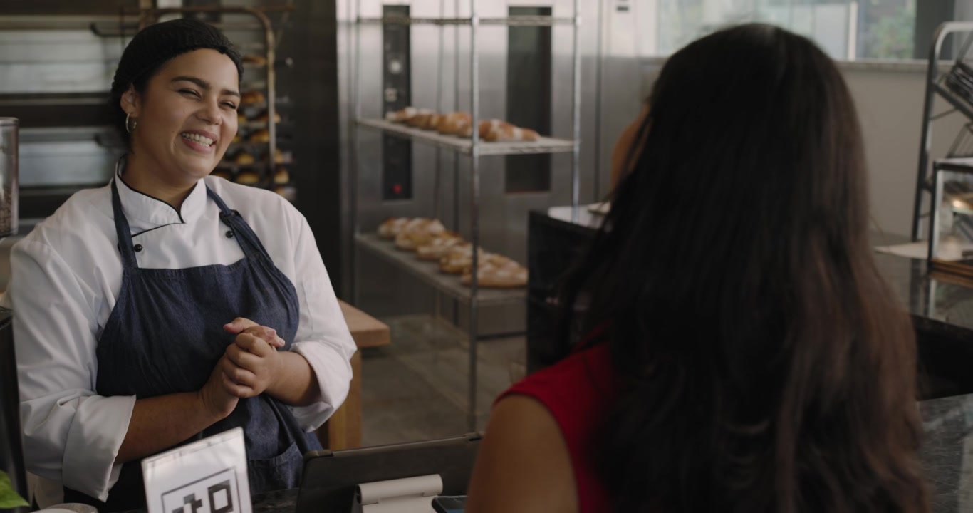 Female baker tapping tablet selecting pastry as customer with phone arriving to buy at counter