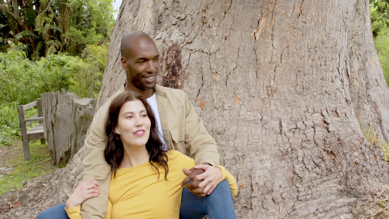 Smiling multiracial couple relaxing under tree, enjoying park and each other's company