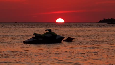 jet ski floats on calm sea surface reflecting red sky at sunset, the sun is setting half down on ocean horizon, summer holidays