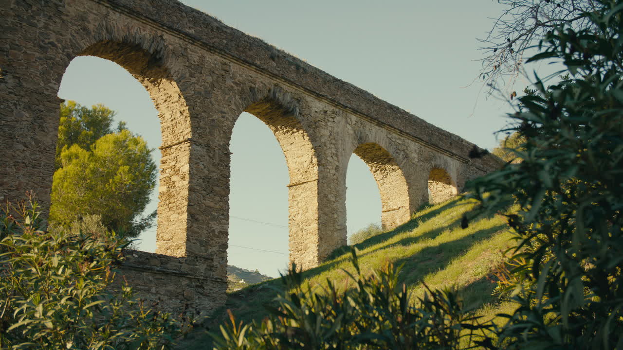 Ancient aqueduct arches glow in golden light as shadows stretch across grassy slope, framed by Mediterranean vegetation